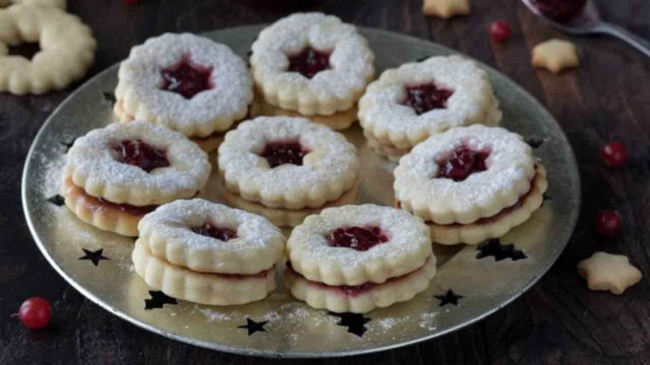 Que tal experimentar uma nova versão dos clássicos biscoitos de Natal? Estes biscoitos leves e saborosos são preparados com massa à base de iogurte, tornando-os uma opção perfeita para quem deseja um toque de leveza, sem abrir mão do sabor e da textura irresistível. A receita é ideal para compartilhar momentos em família, especialmente com as crianças, durante os preparativos natalinos. A substituição parcial da manteiga pelo iogurte cria uma alternativa mais equilibrada, mantendo a maciez e o sabor característico dos biscoitos amanteigados tradicionais. Além disso, são incrivelmente fáceis de preparar e personalizar. Esta receita versátil permite diferentes variações: os biscoitos podem ser guarnecidos com compota, creme de avelã, pistache ou qualquer outro doce festivo à sua escolha. Perfeitos para acompanhar uma xícara de chocolate quente ou para presentear alguém especial, eles certamente serão um sucesso entre jovens e adultos, trazendo um toque original à sua celebração natalina. Como Fazer Biscoitos de Natal com Geléia Ingredientes para 28 biscoitos (ou 14 duplos): 340 g de farinha 125 g de iogurte natural 100 g de açúcar 60 g de óleo de girassol 1 ovo médio 1 colher de chá de fermento em pó (3-4 g) Raspas de ½ limão ½ vagem de baunilha ou 1 colher de chá de extrato de baunilha 1 pitada de sal 150 g de geléia (sabor à sua escolha) Açúcar de confeiteiro, para decorar Modo de Preparo: Misture os ingredientes líquidos: Em uma tigela, bata o ovo com o açúcar e uma pitada de sal até o açúcar dissolver. Acrescente o iogurte, o óleo de girassol, as raspas de limão e o extrato ou sementes de baunilha. Misture suavemente com um batedor até incorporar. Incorpore os secos: Adicione o fermento peneirado e misture com um garfo. Reserve 3 a 4 colheres de sopa de farinha. Aos poucos, acrescente o restante da farinha, amassando primeiro na tigela e depois em uma superfície enfarinhada, até obter uma massa lisa e homogênea. Abra e corte a massa: Pré-aqueça o forno a 170°C (modo estático). Divida a massa em duas partes. Abra uma delas sobre papel manteiga enfarinhado até atingir 3 mm de espessura. Corte formatos de Natal (estrelas, árvores, etc.) ou círculos de 5 cm de diâmetro. Faça aberturas centrais em metade dos círculos usando cortadores menores. Asse os biscoitos: Transfira os biscoitos para uma assadeira forrada com papel manteiga. Asse por 12 minutos, até que a base esteja levemente dourada. Deixe esfriar por alguns minutos na assadeira antes de manusear. Monte os biscoitos: Após esfriar, polvilhe os biscoitos com abertura central com açúcar de confeiteiro. Espalhe meia colher de chá de geléia sobre os biscoitos inteiros e pressione levemente os biscoitos perfurados sobre eles, formando "sanduíches". Dicas Extras: Use geléias de framboesa, morango, damasco ou escolha seu sabor favorito. Reaproveite as sobras de massa para criar mais biscoitos, garantindo o máximo rendimento. Armazene os biscoitos em um recipiente hermético para mantê-los frescos e crocantes por vários dias. Estes biscoitos leves, perfumados e deliciosos são perfeitos para presentear ou para compor sua mesa natalina com charme e sabor. Bom apetite!